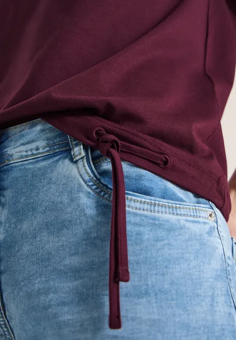 Seitlich geschnürtes bordeauxrotes Shirt kombiniert mit hellblauer Jeans.